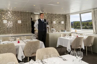 Der Maître in einem eleganten Restaurant auf einem Flusskreuzfahrtschiff von Thurgau Reisen. Der Raum ist geschmackvoll mit weißen Tischtüchern eingedeckt und mit Gläsern und Blumen dekoriert. Große Fenster bieten einen Blick auf die vorbeiziehende Landschaft, während der Maître bereit ist, die Gäste zu empfangen.