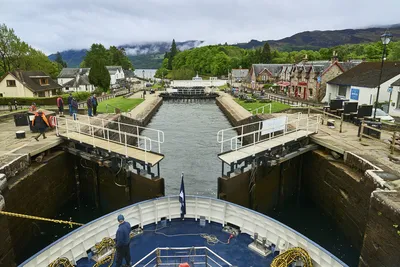 Blick auf eine Schleuse des Caledonian Canal, während ein Flusskreuzfahrtschiff von Thurgau Reisen einfahren möchte. Im Vordergrund ist das Schiff zu sehen, während die Schleusentore geöffnet sind. Im Hintergrund sind charmante Häuser und eine grüne Landschaft sichtbar, die die malerische Umgebung betonen.