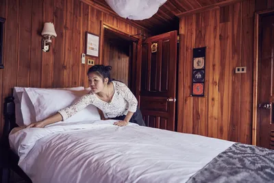 Eine Mitarbeiterin des Housekeeping-Service auf dem Flusskreuzfahrtschiff Mekong Pearl von Thurgau Reisen macht das Bett in einer gemütlichen Kabine. Der Holzinterieur verleiht dem Raum eine warme Atmosphäre, während die Angestellte sorgfältig die Bettwäsche glättet.