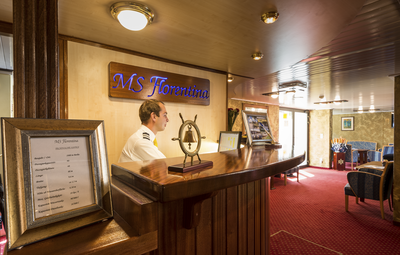 Der Empfangsbereich auf dem Flusskreuzfahrtschiff MS Florentina von Thurgau Reisen, mit dem Hotelmanager hinter dem Empfangstresen. Eine elegante Beschilderung mit dem Namen des Schiffes ist sichtbar, während im Hintergrund gemütliche Sitzgelegenheiten und eine freundliche Atmosphäre auf die Gäste warten.