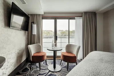Innenansicht einer Kabine des Mitteldecks auf dem Flusskreuzfahrtschiff MS Voyage von Thurgau Reisen. Der Raum ist mit zwei gemütlichen Stühlen und einem kleinen Tisch eingerichtet, vor einem großen Fenster mit Blick auf den Fluss. Die moderne Einrichtung und die sanften Farben schaffen eine entspannte Atmosphäre.