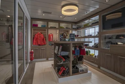 Die Boutique auf dem Flusskreuzfahrtschiff Ocean Albatros von Thurgau Reisen, die eine Auswahl an Kleidung und Accessoires anbietet. Regale aus Holz präsentieren farbenfrohe Jacken und Souvenirs, während eine moderne Beleuchtung dem Raum eine einladende Atmosphäre verleiht.
