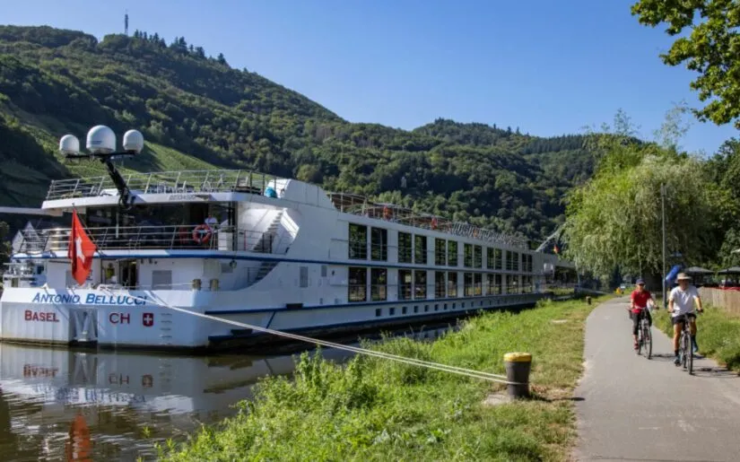 Kreuzfahrtschiff Thurgau Reisen Antonio Bellucci, das an einer Anlegestelle vertäut wurde. Daneben ist ein Fußweg und bewaldete Berge zu sehen