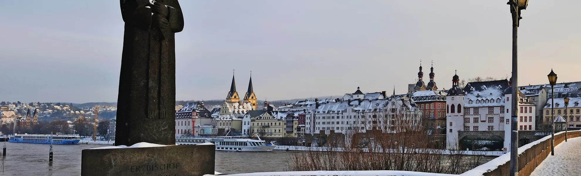 Blick auf Koblenz mit verschneiter Statue und Gebäuden entlang des Rheins im Winter.